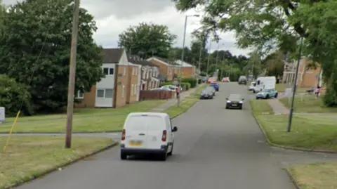 Generic Google image of a white van driving down a residential road, with other vehicles in the background. There are lamp-posts and houses along the street.