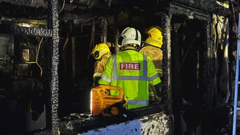 Shropshire Fire and Rescue Service Firefighters are standing inside a charred, burnt-out room. Everything is black after the fire and the firefighters are wearing hi-vis jackets.