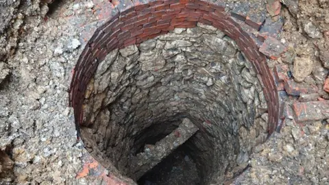 A circular well shaft lined with worn stones, looking down towards a stone cross beam and rubble at the base. Towards the top of the well are about eight layers of red bricks. More stones, rubble and bricks surround the well.