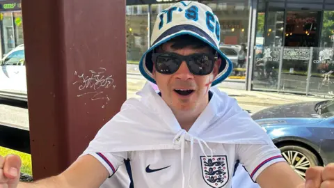 Three Lions superfan Andy Taylor. He is wearing an England shirt and bucket hat, and has a St George's flag with him.
