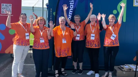 Group of six woman standing cheering, wearing orange tops