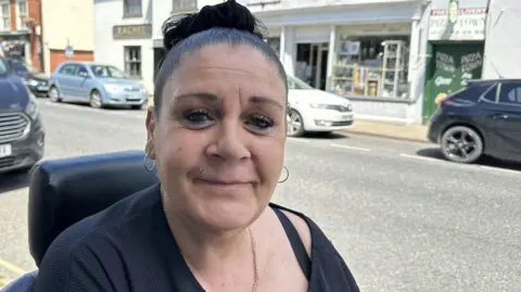Ben Parker/BBC A woman with dark hair, she is looking at the camera and wearing a black top. In he background are cars parked on the street.