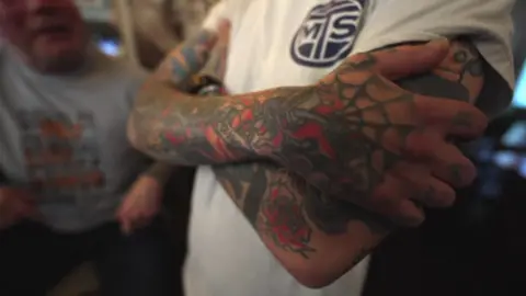 A close up of a tattooed arm. The person is wearing a white t-shirt. Their tattoo uses red and green ink. 