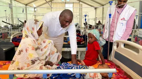 David Lamy Leaning over a bed in a white shirt a mother sits with a baby and child at an MSF clinic in Chad. An MSF medic is standing nearby