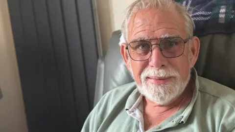 George King/BBC A head and shoulders image of Graham West. He is wearing glasses and a green polo shirt and sitting in a leather chair. He is looking into the camera.