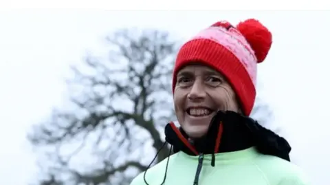 Prem Management A woman with a red knitted bobble hat and green and black jacket stands outside. The outline of a tree can be seen behind her.