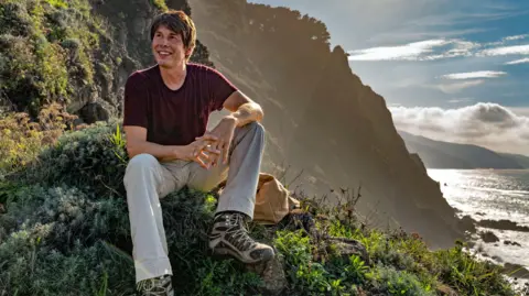 BBC Studios/Nick Gavin Il professor Brian Cox siede su una collina coperta di alberi, con il mare sul lato destro dell'immagine. Indossa una camicia bordeaux, pantaloni grigi e scarpe da passeggio. Sorride mentre guarda in lontananza. Il sole splende luminoso sullo sfondo.