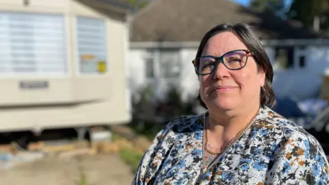 BBC Phoenix Graham stands successful  the driveway of her location  successful  Ruskington, Lincolnshire. She is wearing a floral blouse and dark-rimmed glasses. In the inheritance  stands a cream-coloured static caravan, adjacent  to a white-walled bungalow.