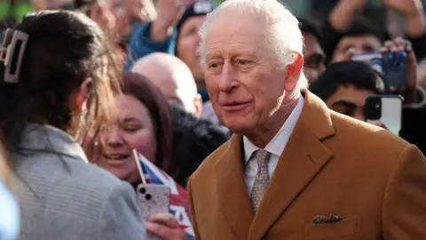 King Charles talking to a woman who has her back to the camera. A crowd of people can be seen behind him.