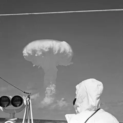 Mirrorpix via Getty Images A person in the foreground seen from behind, wearing a white hood and jacket, looks out to sea as mushroom cloud rises into the air.