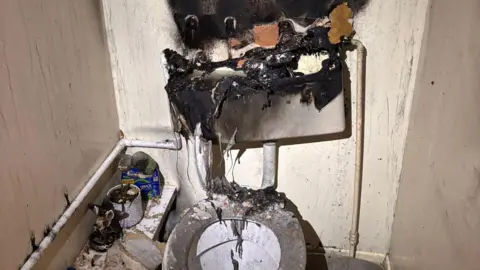 A burnt out toilet, with burnt wall, a burnt cistern and burnt items to the left. The room is full of soot. 