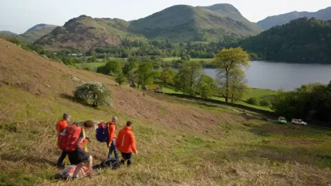 BBC Four people in orange jackets walk down a grassy hill pulling an empty stretcher. They are heading towards two white Land Rovers with orange and green markings, with a lake and hills beyond