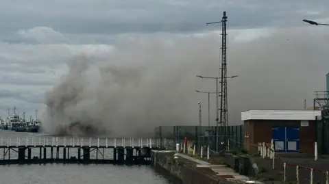 Amber Gash/BBC A large plume of grey smoke envelops a large cargo ship. It is visible behind a set of lock gates.