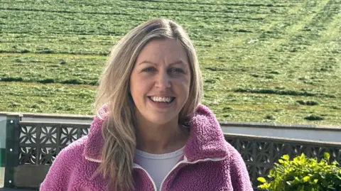 Dr Eimear Rooney Dr Eimear Rooney is standing in front of a freshly cut field, smiling. Her hair is blonde and she is wearing a pink, fleecy jacket over a white t-shirt. 