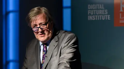 Richard Eaton, Kings College London Stephen Fry, standing at a lectern in a suit and tie