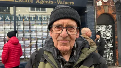 Derek wears glasses and a black knitted hat and is standing in front of the estate agents.  He smiles for the camera.