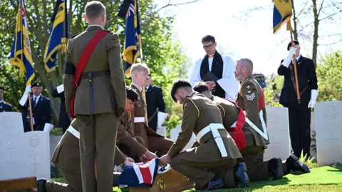 Ministry of Defence/Crown Copyright/PA Wire The coffin of Pte Reginald Joseph Blake being laid to rest at a burial service at the Commonwealth War Graves Commission's Loos British Cemetery Extension, on Wednesday 22 April 2026. Six soldiers in the uniform of the Royal Anglian Regiment are kneeling on either side of the grave as they fold a union jack over his coffin. A seventh soldier is standing to attention. There are white Commonwealth War Gfaves Commisison headstones, a chaplain in a white and men holding flags. 
