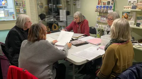 Five women sit around a table. They are all wearing jumpers and are holding lists of names and dates from the gravestones. 