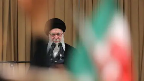Ayatollah Ali Khamenei speaks into microphones with a blurred Iranian flag positioned in front of him near the camera lens.