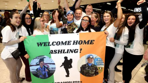 PA Media A group of people surround Pte Ashling Keogh. There are 12 of them and they are all standing behind a banner of the Irish flag with two pictures of Pte Keogh on duty with the peacekeeping force. The top of the flag has Welcome Home Ashling! at the top. Everyone is looking jubilant and they are either cheering or raising their hands in the air.