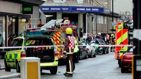 Getty Images Emergency response scene outside Edgware Road Station with multiple fire engines, an ambulance, and emergency personnel. A firefighter in a yellow helmet and reflective gear stands near the vehicles. The area is cordoned off with tape, and there are several onlookers in the background.