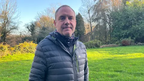 Clare Worden/BBC John McKean looks serious at the camera as he is photographed outside in a green space. He is wearing a blue puffer jacket with a navy fleece beneath. He has short hair and stubble. 