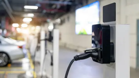 Getty Images A close-up view of an electric vehicle charging station in an indoor parking area. The foreground shows a black charging connector plugged into a sleek black charging unit mounted on a white pillar. In the background, several parked cars and additional charging stations are visible.