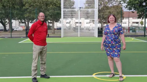  Will Davies on the left in a red jumper, brown trousers and black trousers smiling at the camera and Joanne Kerr on the right in a blue patterned dress smiling at the camera. They are standing on the new play zone area in Tile Hill