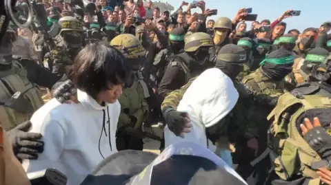 BBC Two Thais taken hostage by Hamas are freed in Khan Yunis on January 30, 2025 - grab from video by BBC Gaza freelancer. Two men wearing white hoodies are surrounded by masked guards and people trying to take their pictures with their mobile phones