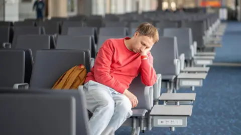 Getty Images Man asleep on a chair in airport departure lounge. He is resting on his hand and all the other chairs around him are empty.