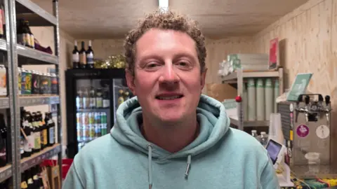 Paul Moseley/BBC Toby Westgarth is standing inside the shipping container which is home to Sir Toby's Beers. He is looking at the camera and smiling and is wearing a light blue hoodie. Behind him are shelves of craft beers in cans and glass bottles and there is a refrigerator behind him. 