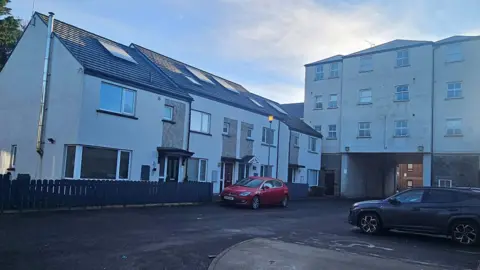 Charlene Mongan A row of white houses and and an apartment complex. 
Two cars are parked outside of two homes in car parking spaces. 