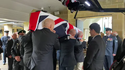 The image shows people carrying a coffin which has a Union Jack flag draped over the top of it. There are people watching as it's carried into the crematorium. 