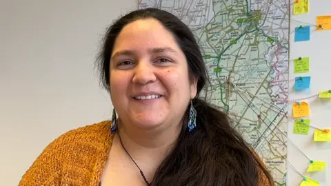 Tom Jackson/ BBC Ms Avilez is wearing a yellow knitted cardigan and long blue, white and green earrings. She has long brown hair, in a ponytail over her shoulder, brown eyes and is smiling at the camera. Behind her is a map on the wall with string and notes pointing to, and sharing comments, about the town of March. 