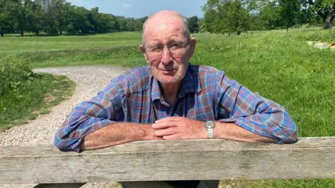 Ron Wood is a 78 year old man wearing a blue and brown shirt leaning on a wooden gate. there is a path and some fields behind with a number of trees on. He has thinning grey hair and glasses 