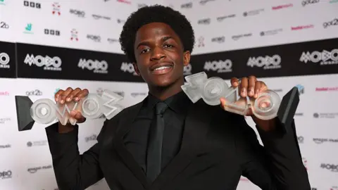 Getty Images DC3 poses with the award for Best Gospel Act and the Best Newcomer award inside the winners' room during the 2026 MOBO Awards. He is wearing a black suit with a black shirt and black tie. 
