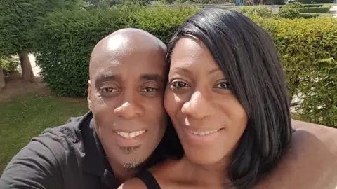 Pauline Reid A man who is bald, smiling at the camera for a selfie. He is wearing a black t-shirt and has a short grey and black goatee. He has his arm around a woman who has a black bob. They are in a garden with green grass, trees and bushes behind them