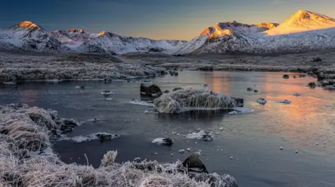 Andrew Briggs A frozen loch is surrounded by frosted grass with mountains in the background. The water is frozen with a number of rocks poking through the ice. At the centre of the loch is a small island covered in frozen grass. In the background, a snowy mountain range with six peaks covered in snow and dark-coloured land can be seen. Orange light from the morning sun covers two at the far end and the sky is dark blue, fading to light blue and orange.