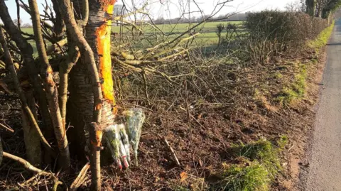 The side of a rural road showing bunches of flowers propped up against a tree. The hedge to the right, between the road and a field, appears to have been damaged.