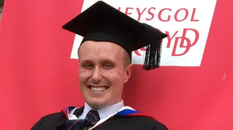 Jack Tagg smiles at the camera. He is sat in an oversized deck hair and is wearing a graduation gown and hat.