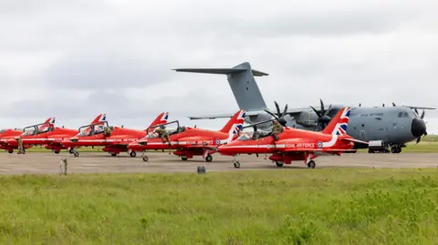 MOD Five red arrow hawk jets on a runway, with a larger grey aircraft behind the reds.