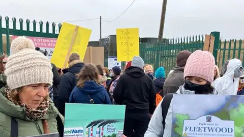 Demonstrators with woolly hats and face coverings holding placards with colourful cartoons outside the tip. Signs read Toxic Stop The Stink and Welcome to Fleetwood - Twinned with Stinkstown, Dumperville. 