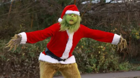 Nuneaton Grinch/Kevin Hargrave  A man dressed as the Grinch standing in front of a hedge. He has his arms outstretched. 