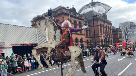 A man wearing brown trousers and a purple coat is sitting on top of an effigy of a horse. A man cycling next to him has an umbrella. Others performers are in the background. A crowd is watching. 