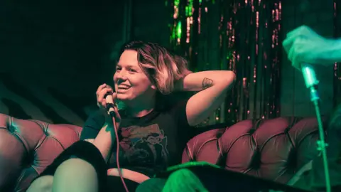 Connor Madehell Shannon Davies seated on a tufted leather sofa holding a microphone during a live recording or interview, with colorful green and pink stage lighting and shimmering backdrop decorations. Another microphone is visible in the foreground, creating an intimate event or podcast setting.
