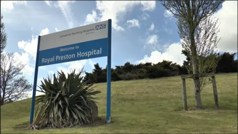 A blue and white Royal Preston Hospital sign with a large green plant underneath it, surrounded by lawn grass