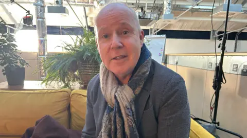 balding man with jacket and scarf sitting on yellow sofa with plants, technical rigs and cameras in the background