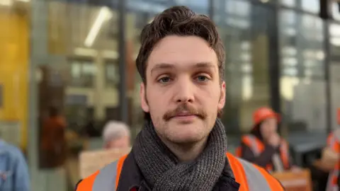 George Carden/BBC Close up of young man looking to camera outdoors with moustache and wearing scarf and orange hi vis jacket