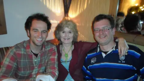 A woman with curly blonde hair, wearing a blue top and red cardigan. On either side of her is a man - the one on the left has dark hair and is wearing a red and green check shirt, while the one on the left is wearing a blue and navy striped rugby shirt and has dark hair and glasses. All three people are smiling and looking at the camera. They are sat at a table with glasses on it. Behind them is a wall with a mirror hanging.