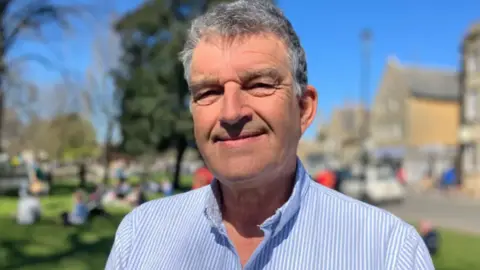 A picture of Andrew Lund-Yates who is smiling directly at the camera. He has grey hair and is wearing a blue and white shirt, open at the neck. He is standing outdoors on a sunny blue-sky day and buildings and people can be seen in the background. 